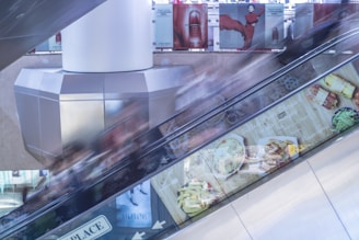 An escalator is situated in a shopping mall with motion blur caused by moving people. Advertisements line the side of the escalator, featuring images of food and beauty products. The scene includes a structural column in the middle and a clean, modern aesthetic with metallic and glass elements.