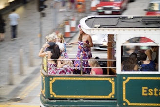 A group of people wearing colorful outfits are enjoying a ride on an open-top vintage tram. The scene appears to be vibrant and lively, with some individuals standing and others sitting casually. The background includes blurred images of other vehicles and people, suggesting motion and a busy urban environment.
