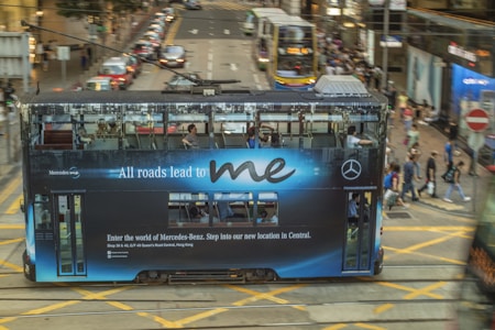 A streetcar displaying an advertisement for Mercedes-Benz drives down a busy city street. The ad message, 'All roads lead to me,' is prominently visible on the side of the tram. Pedestrians cross the street in the background while other vehicles and city buildings are visible, conveying a bustling urban atmosphere.