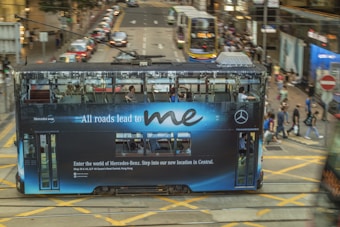 A streetcar displaying an advertisement for Mercedes-Benz drives down a busy city street. The ad message, 'All roads lead to me,' is prominently visible on the side of the tram. Pedestrians cross the street in the background while other vehicles and city buildings are visible, conveying a bustling urban atmosphere.