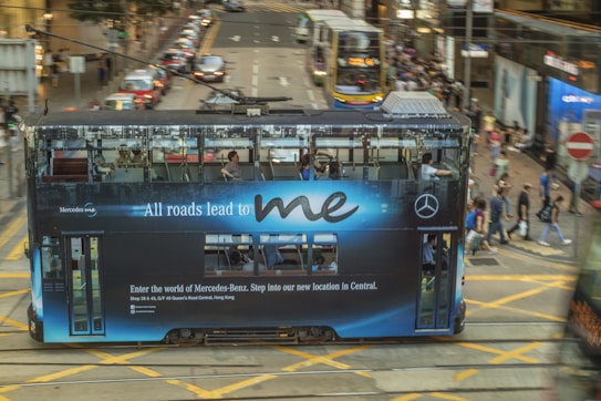 A streetcar displaying an advertisement for Mercedes-Benz drives down a busy city street. The ad message, 'All roads lead to me,' is prominently visible on the side of the tram. Pedestrians cross the street in the background while other vehicles and city buildings are visible, conveying a bustling urban atmosphere.