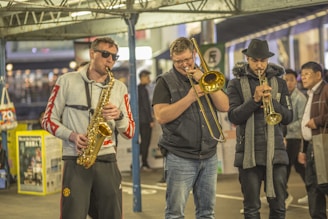 Three musicians perform in a public space, each playing a different brass instrument. The first musician, wearing sunglasses and a grey hoodie with red sleeves, plays a saxophone. The second musician, in a black puffer vest, plays a trombone. The third musician is wearing a black fedora and scarf and plays a trumpet. Pedestrians in the background, hinting at a busy or bustling environment.