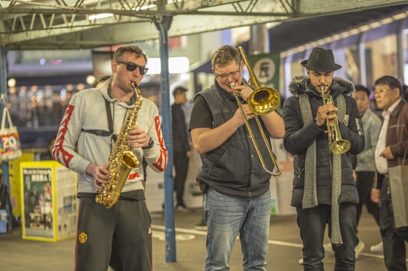 Three musicians perform in a public space, each playing a different brass instrument. The first musician, wearing sunglasses and a grey hoodie with red sleeves, plays a saxophone. The second musician, in a black puffer vest, plays a trombone. The third musician is wearing a black fedora and scarf and plays a trumpet. Pedestrians in the background, hinting at a busy or bustling environment.
