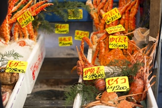 A vibrant seafood market display featuring a variety of crabs. The crabs are arranged on ice within styrofoam boxes. Bright yellow price tags with red and black Japanese writing are placed above the crabs, indicating their types and prices. Green pine branches are used decoratively around the seafood.