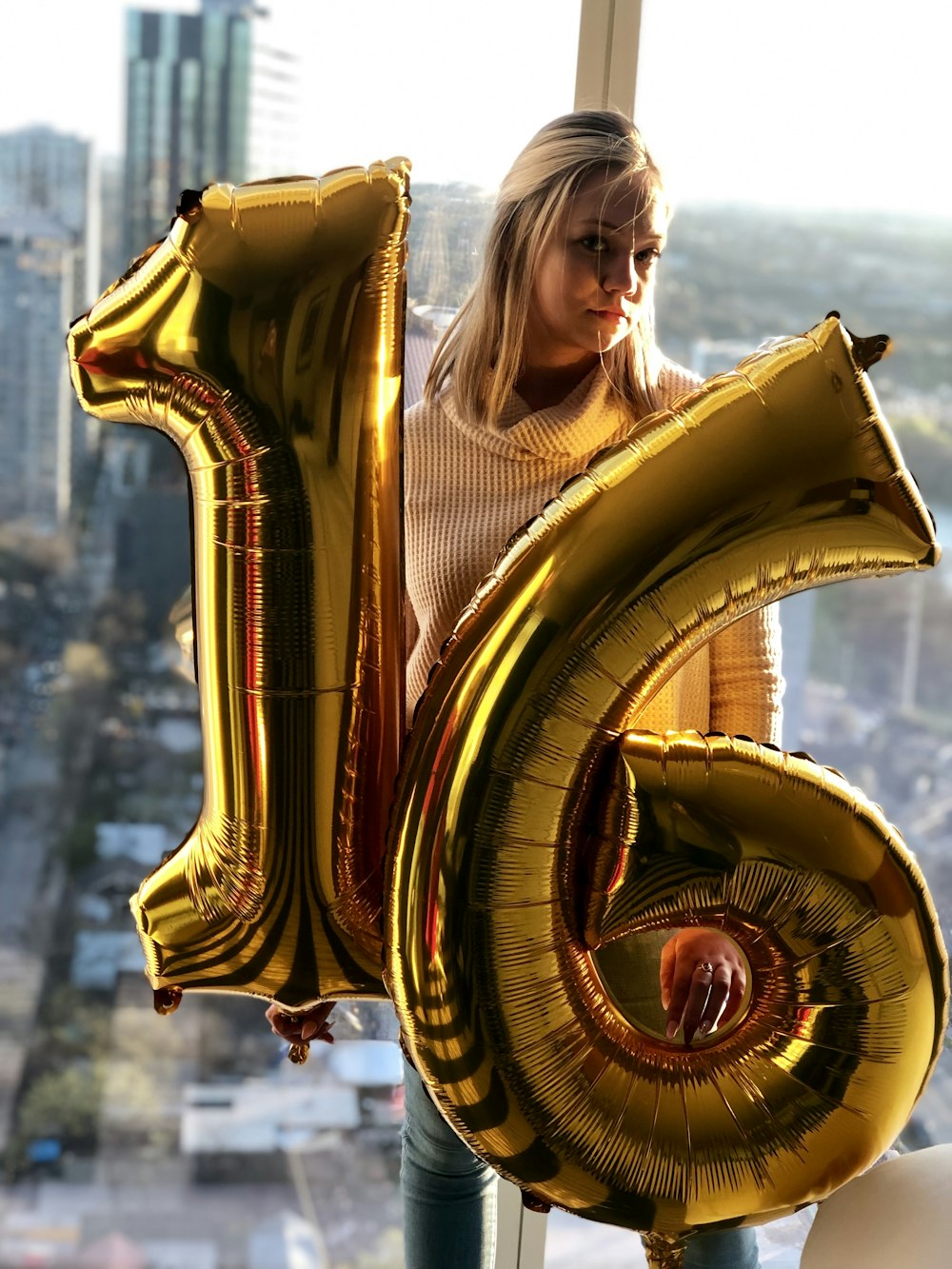 woman holding gold number 16 balloons standing beside glass window