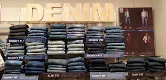 Stacks of jeans organized by fit type are displayed on shelves. Labels indicate styles such as 'Skinny Fit' and 'Slim Fit.' An illuminated 'DENIM' sign is prominently positioned above the display. A poster featuring models wearing various styles of denim is featured on the right side of the display.
