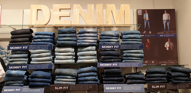 Stacks of jeans organized by fit type are displayed on shelves. Labels indicate styles such as 'Skinny Fit' and 'Slim Fit.' An illuminated 'DENIM' sign is prominently positioned above the display. A poster featuring models wearing various styles of denim is featured on the right side of the display.