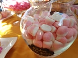A glass jar filled with pink and white marshmallow-like candies is placed on a table. The table is covered with a yellow cloth. In the background, there are more candies in a glass bowl and some on a plate.