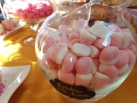 A glass jar filled with pink and white marshmallow-like candies is placed on a table. The table is covered with a yellow cloth. In the background, there are more candies in a glass bowl and some on a plate.