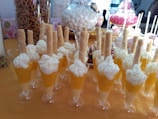 A table filled with desserts in small glasses, each topped with whipped cream and a wafer stick. The background shows an assortment of snacks, including pretzels in a jar and various candies in glass containers.