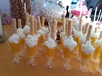 A table filled with desserts in small glasses, each topped with whipped cream and a wafer stick. The background shows an assortment of snacks, including pretzels in a jar and various candies in glass containers.