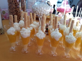 A table filled with desserts in small glasses, each topped with whipped cream and a wafer stick. The background shows an assortment of snacks, including pretzels in a jar and various candies in glass containers.