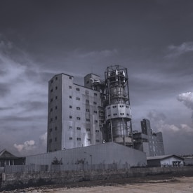 A large industrial building with multiple levels and sections, featuring metal cladding and numerous windows. The structure includes a tall cylindrical tower and a rectangular main building. The sky is overcast with clouds, contributing to a moody ambiance.