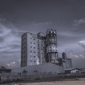 A large industrial building with multiple levels and sections, featuring metal cladding and numerous windows. The structure includes a tall cylindrical tower and a rectangular main building. The sky is overcast with clouds, contributing to a moody ambiance.