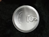 A metal pull-tab can lid is prominently featured, with engraved instructions that read 'LIFT RING PULL STRAIGHT UP'. The lid appears shiny and reflective, indicating it is made of aluminum or similar material. The background is a textured, dark surface.