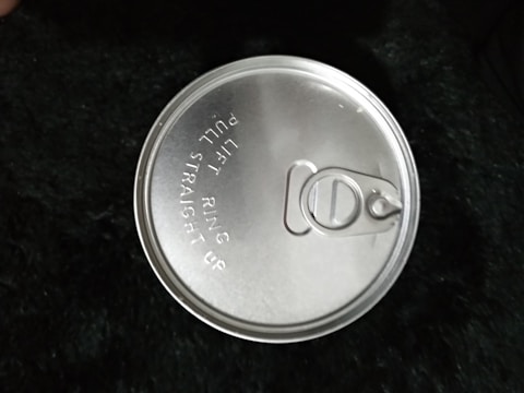 A metal pull-tab can lid is prominently featured, with engraved instructions that read 'LIFT RING PULL STRAIGHT UP'. The lid appears shiny and reflective, indicating it is made of aluminum or similar material. The background is a textured, dark surface.