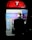 A person is using a public payphone booth that offers free Wi-Fi for Telstra customers. The booth is illuminated with a bright red top featuring a large letter 'T'. Inside, information panels and the payphone are visible. The scene is set against a dark background, highlighting the booth and user.