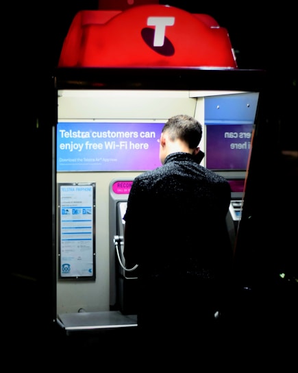 A person is using a public payphone booth that offers free Wi-Fi for Telstra customers. The booth is illuminated with a bright red top featuring a large letter 'T'. Inside, information panels and the payphone are visible. The scene is set against a dark background, highlighting the booth and user.