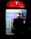 A person is using a public payphone booth that offers free Wi-Fi for Telstra customers. The booth is illuminated with a bright red top featuring a large letter 'T'. Inside, information panels and the payphone are visible. The scene is set against a dark background, highlighting the booth and user.