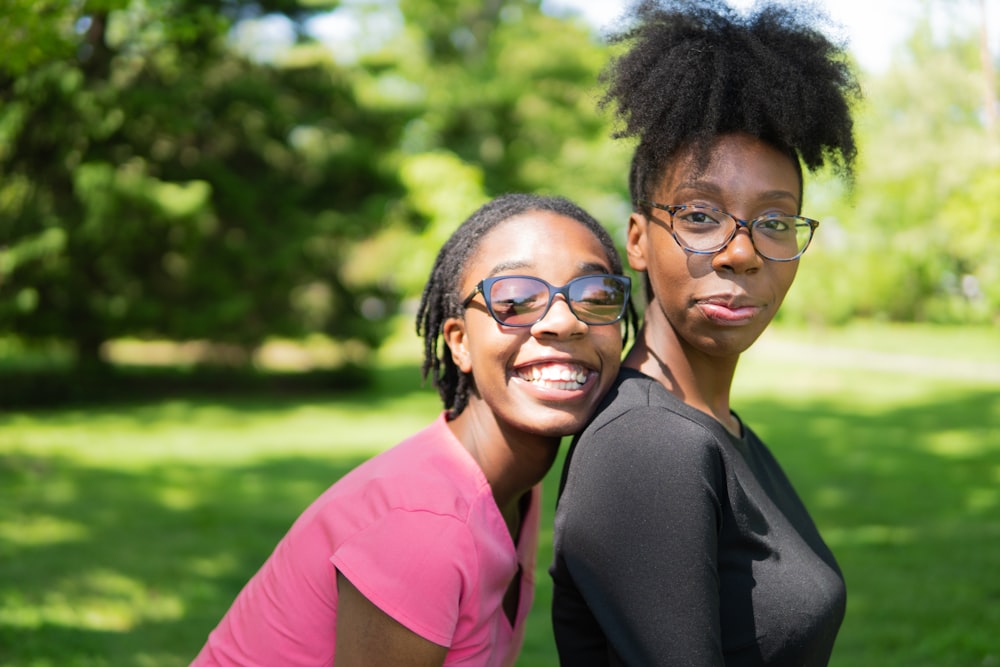 smiling woman at the back on another woman standing on green field surrounded with tall and green trees