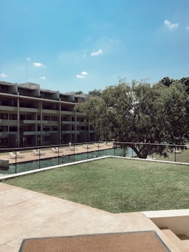 A serene outdoor setting featuring a modern, multi-story building with balconies overlooking a swimming pool area. The pool is surrounded by sunbeds, and there is a large leafy tree in the foreground. The sky is clear with a few white clouds, and the grass is neatly maintained.