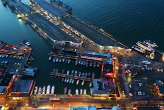 An aerial view of a waterfront area featuring multiple docks with boats and yachts moored alongside them. The image includes several large pier structures extending into the water and various buildings illuminated with artificial lights, giving the scene a vibrant night-time appearance. The water displays a dark blue hue, contrasting with the bright, warm lights from the structures and streets. Additional boats are arranged neatly along the docks, and a few streets and parking areas are visible, indicating a bustling marina setting.