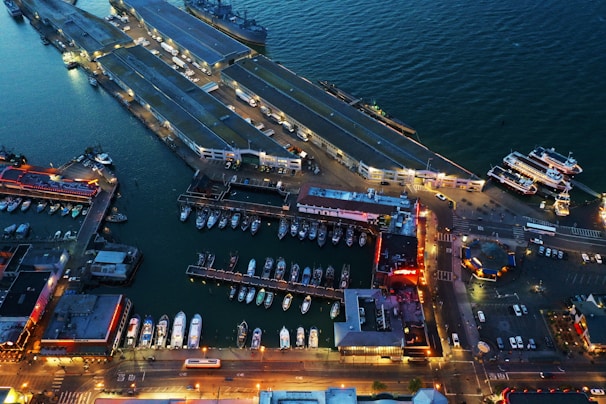 An aerial view of a waterfront area featuring multiple docks with boats and yachts moored alongside them. The image includes several large pier structures extending into the water and various buildings illuminated with artificial lights, giving the scene a vibrant night-time appearance. The water displays a dark blue hue, contrasting with the bright, warm lights from the structures and streets. Additional boats are arranged neatly along the docks, and a few streets and parking areas are visible, indicating a bustling marina setting.