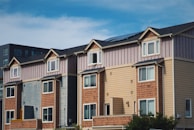 A row of townhouses or apartment buildings with multiple stories and gabled roofs. The exterior walls display a mix of colors and materials, including shades of brown, beige, and grey. Large windows are visible, and there is a clear blue sky above.