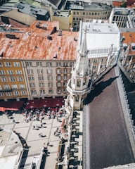 Aerial view of a city square featuring historical architecture, including a spire and multi-story buildings with red rooftops. A number of people are gathered at outdoor cafés with red umbrellas, along a patterned stone pavement.