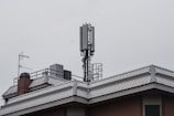 A rooftop features a mobile phone antenna tower and a satellite dish amidst other rooftop equipment. The sky is overcast, giving a grayish tone to the scene.