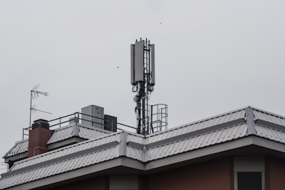 A rooftop features a mobile phone antenna tower and a satellite dish amidst other rooftop equipment. The sky is overcast, giving a grayish tone to the scene.