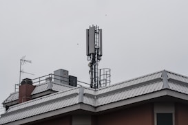 A rooftop features a mobile phone antenna tower and a satellite dish amidst other rooftop equipment. The sky is overcast, giving a grayish tone to the scene.