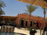 An outdoor seating area with metal chairs and a glass table is set under a wooden pergola. The background features a building with an architectural style that incorporates arched windows framed in red and green, with a peach-colored facade. Several palm trees are planted along the open space, lending a tropical atmosphere.