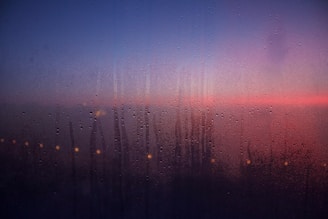 Condensation covers a glass pane with delicate water droplets, forming a misty surface. Through the moist glass, a serene sunset displays gradients of blue, purple, and pink hues in the sky, while a line of blurred warm lights suggests distant city lights or street lamps.