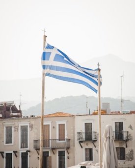 Two flagpoles hold Greek flags with blue and white stripes and a cross on a clear day. Historical buildings with balconies and aged facades are visible in the background with a hazy mountain range.