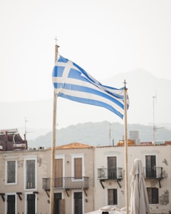Two flagpoles hold Greek flags with blue and white stripes and a cross on a clear day. Historical buildings with balconies and aged facades are visible in the background with a hazy mountain range.