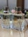 Two elegant glass perfume bottles with gold lids are standing side by side on a wooden surface. Each bottle is adorned with a leather strap around the neck, one in white and the other in tan. A small pink rose and some greenery serve as a decorative background, adding a touch of delicacy to the scene.