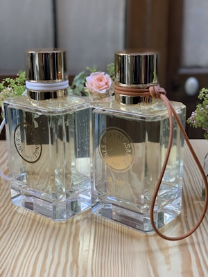 Two elegant glass perfume bottles with gold lids are standing side by side on a wooden surface. Each bottle is adorned with a leather strap around the neck, one in white and the other in tan. A small pink rose and some greenery serve as a decorative background, adding a touch of delicacy to the scene.