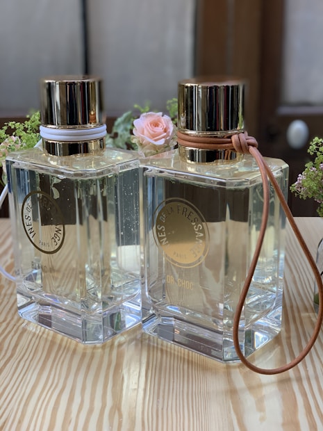 Two elegant glass perfume bottles with gold lids are standing side by side on a wooden surface. Each bottle is adorned with a leather strap around the neck, one in white and the other in tan. A small pink rose and some greenery serve as a decorative background, adding a touch of delicacy to the scene.