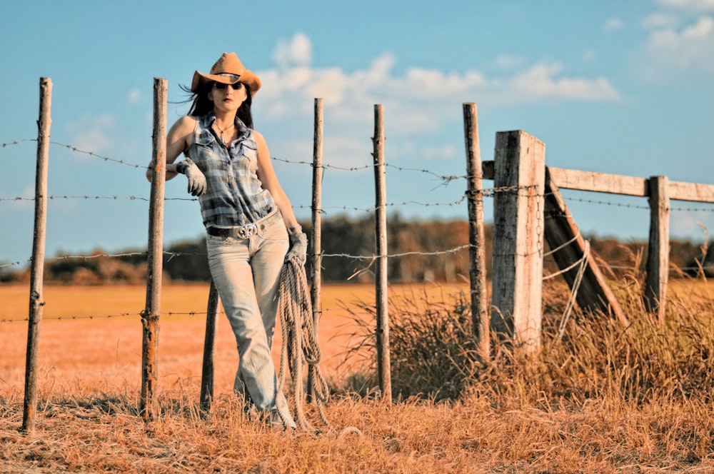 Mujer en blusa abotonada sin mangas y sombrero de vaquero apoyada en la cerca de alambre de púas