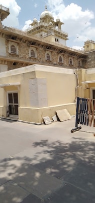 A historic building with ornate architectural details, featuring arched windows and decorative railings, topped with domes and spires. The structure is made of stone and plaster, giving it a traditional appearance. In the foreground, there are paving stones, shadows from trees, and some minor construction materials.