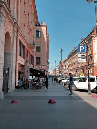 A city street lined with buildings featuring outdoor cafes where people are sitting at tables. Cars are parked along the street next to a sidewalk, and several pedestrians are walking. Overhead, a clear blue sky is visible. A prominent parking sign is displayed on the right with a 20-meter restriction.
