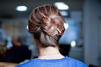 The image depicts the back of a person's head with hair styled into a neat bun, secured with a hair stick. The hair is brown with lighter highlights. The person is wearing a blue top and appears to be indoors, with a blurry background of what seems like an office or workspace.