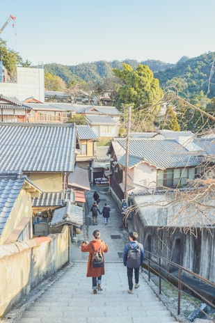 A scenic view of a traditional Japanese street with people walking down a stone stairway. The roofs of the houses are tiled with a traditional design, surrounded by lush green hills in the background. The sunlight casts a soft glow over the area, creating a peaceful atmosphere.