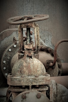 A rusty metal valve is prominently featured, showcasing signs of age and corrosion. The background reveals other industrial components, with bolts and pipes visible. The surroundings appear weathered, adding to the industrial and aged look.