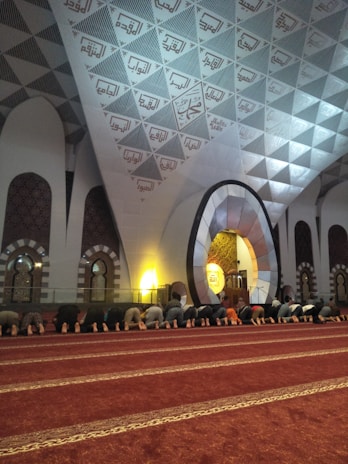 A group of people are engaged in prayer, kneeling on a red carpet with ornate gold patterns. The architectural setting features large arches and intricate geometric designs on the walls and ceiling, with Arabic calligraphy present. Soft, ambient lighting adds to a serene atmosphere.