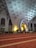 A group of people are engaged in prayer, kneeling on a red carpet with ornate gold patterns. The architectural setting features large arches and intricate geometric designs on the walls and ceiling, with Arabic calligraphy present. Soft, ambient lighting adds to a serene atmosphere.