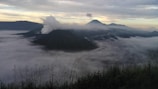 A scenic view of mountains partially covered by a thick layer of clouds. Tall grasses are visible in the foreground, while a plume of smoke rises from one of the volcanic peaks. The sky is a mixture of soft pastel colors as the sun begins to rise or set, casting a serene atmosphere over the landscape.