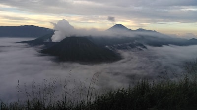 A scenic view of mountains partially covered by a thick layer of clouds. Tall grasses are visible in the foreground, while a plume of smoke rises from one of the volcanic peaks. The sky is a mixture of soft pastel colors as the sun begins to rise or set, casting a serene atmosphere over the landscape.