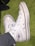 A pair of white high-top sneakers with a rubber sole and a red and blue logo on the side. The sneakers have white laces and silver eyelets. They are being worn by someone wearing grey pants and are photographed on a green textured surface.